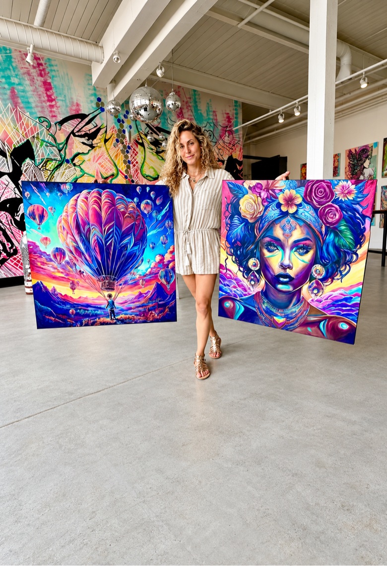 Jessica in her gallery holding two large paintings, a hot-air-balloon scene and a portrait crowned with roses